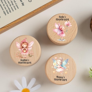 May include: Three wooden tooth boxes, each decorated with a fairy illustration. The boxes feature different fairy designs in pastel colours, with names such as "Sophia's," "Bella's," and "Nancy's" printed below. The boxes are round and made of light-coloured wood.