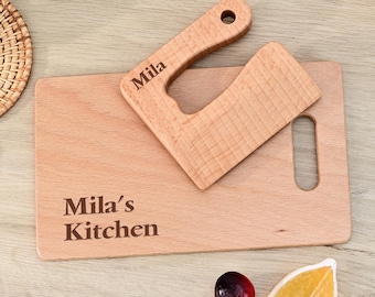May include: A wooden cutting board and cleaver set. The cutting board is rectangular with the text "Mila's Kitchen" engraved on it. A small cleaver with the name "Mila" is on top. A cherry and a lemon slice are next to the board.