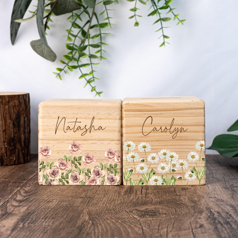 May include: Two wooden piggy banks with floral designs. The piggy bank on the left has a pink rose design and the name "Natasha" engraved on it. The piggy bank on the right has a white daisy design and the name "Carolyn" engraved on it.