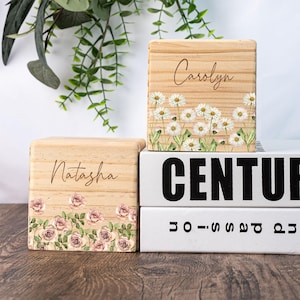 May include: Two wooden blocks with floral designs and personalized names. The top block has a white daisy design and the name "Carolyn" engraved on it. The bottom block has a pink rose design and the name "Natasha" engraved on it.