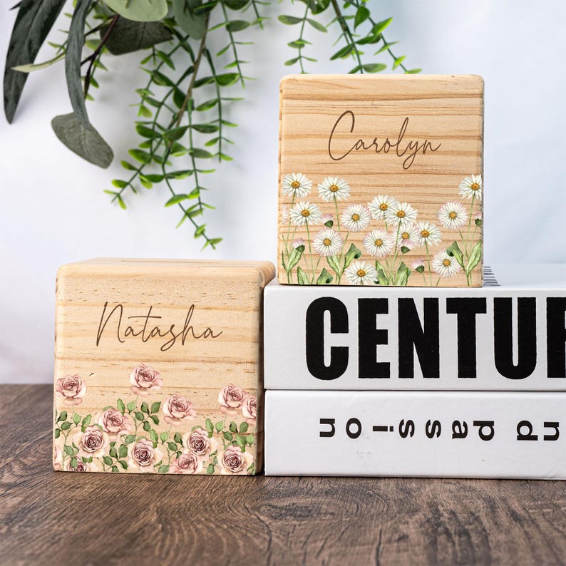 May include: Two wooden blocks with floral designs and personalized names. The top block has a white daisy design and the name "Carolyn" engraved on it. The bottom block has a pink rose design and the name "Natasha" engraved on it.