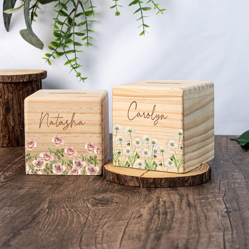 May include: Two wooden piggy banks with floral designs. One has a pink rose design and says "Natasha" on the front. The other has a white daisy design and says "Carolyn" on the front.