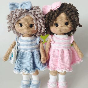 May include: Two handmade crochet dolls. One doll wears a blue dress with a matching bow, shoes, and a small purse. The other doll wears a pink dress with a matching bow and shoes. Both dolls have curly hair and are wearing white socks.