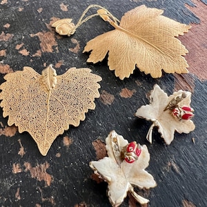 May include: A collection of four leaf-shaped pins including one large gold-toned textured maple leaf with a hanging tag, one gold-toned filigree leaf, and two smaller white enameled maple leaves each featuring a small red ladybug accent.