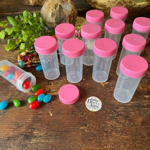May include: A collection of clear plastic jars with pink screw-on lids. Some jars are upright, while others are scattered on a wooden surface. Colorful jelly beans are spilled from one jar. A sticker reads "#3314 Deco Jars.com".