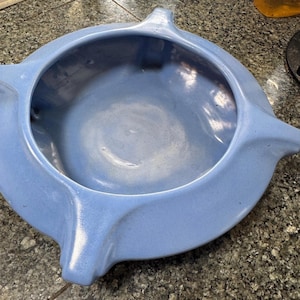 May include: A light blue ceramic bowl with a smooth, glossy finish. The bowl features a wide, flat rim that extends outward into four distinct, evenly spaced points, creating a star-like silhouette around the central circular basin.