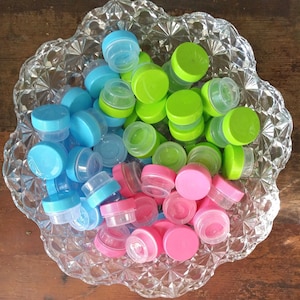 May include: A clear, decorative glass bowl filled with small, colorful plastic containers. The containers have blue, green, and pink lids. The bowl sits on a wooden surface.