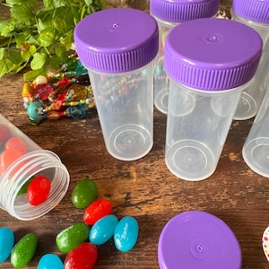 May include: Clear plastic containers with purple screw-on lids. Several containers are upright, while one is tipped over, spilling colorful candies. The candies are red, green, and blue, with some labeled "Starburst". A beaded bracelet and greenery are in the background.