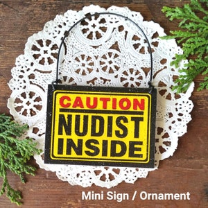 May include: A black and yellow sign with the text "CAUTION NUDIST INSIDE" on a white doily. The sign is attached to a black metal hanger. Mini Sign / Ornament.
