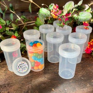 May include: A collection of clear plastic jars with white screw-on lids. One jar is filled with colorful jelly beans. The jars are arranged on a dark wooden surface, with greenery and flowers in the background. The lid of one jar has a sticker with the text "#4314 Deco Jars .com".
