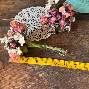 May include: A bouquet of small, colorful artificial flowers with pink, purple, and yellow blooms, tied with green tape. The bouquet rests on a white lace doily. A yellow measuring tape shows the bouquet is approximately 6 inches long.