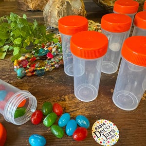 May include: Clear plastic jars with bright orange lids are arranged on a wooden surface. Colorful candies are scattered around a jar that has been tipped over. A sticker with the text "Deco Jars" is also visible.