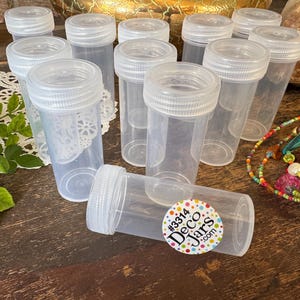 May include: A collection of clear plastic jars with white screw-on lids. One jar lies horizontally, displaying a colorful sticker with text. The jars are arranged on a dark wooden surface, with decorative elements in the background.