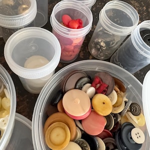 May include: A collection of assorted buttons in various shapes, sizes, and colors, including shades of beige, pink, brown, black, and white, stored in several clear plastic containers with screw-top lids, organized by color and style.