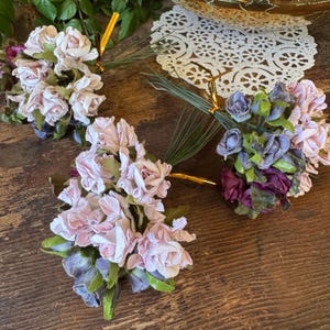May include: Three bouquets of artificial flowers in shades of pink, purple, and green. The flowers are arranged in small clusters and tied with gold wire. The bouquets are set on a wooden surface.