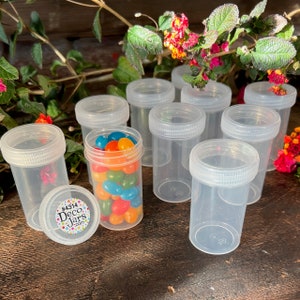 May include: Clear plastic jars with white lids, some filled with colorful jelly beans. The jars are arranged on a wooden surface, with a sticker that reads "#4314 Deco Jars .com". Decorative flowers and greenery are in the background.