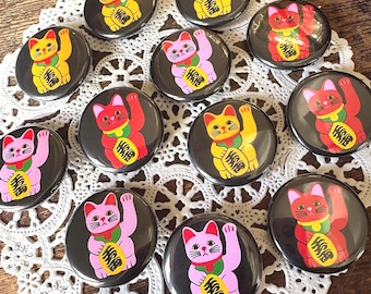 May include: A collection of ten circular pin-back buttons featuring illustrations of beckoning cats in yellow, pink, and red. Each cat is depicted with a green collar and a yellow coin marked with black Japanese kanji characters.