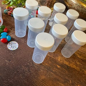 May include: A collection of clear plastic jars with white screw-on lids. The jars are cylindrical and arranged on a dark wooden surface. A small sticker with text is visible. The jars are empty, suggesting they are for storage or display of small items.