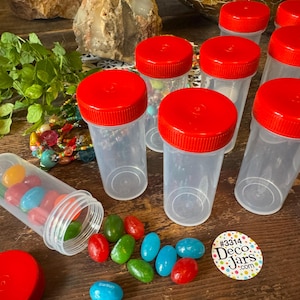 May include: Clear plastic jars with red lids are arranged on a wooden surface, some filled with colorful candies. A few candies are scattered around the jars. A sticker with the text "#3314 Deco Jars .com" is also visible.