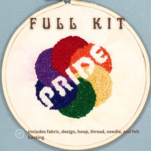 May include: A white embroidery hoop with the word "FULL KIT" printed at the top. The hoop contains a rainbow-colored embroidery design that says "PRIDE". The description below the hoop says "Includes fabric, design, hoop, thread, needle, and felt backing."