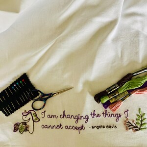 May include: White fabric with embroidery tools and threads. The fabric is embroidered with a unicorn and the quote "I am changing the things I cannot accept" by Angela Davis. Includes needles and scissors.