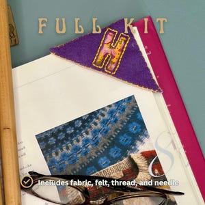 May include: A purple felt bookmark with a felt letter H in the center. The bookmark is on top of a book with a close-up image of a knitted sweater. The text "FULLKIT" is above the bookmark. The text "Includes fabric, felt, thread, and needle" is below the bookmark.