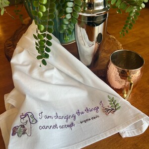 May include: White cotton tea towel with embroidered unicorn and floral design. The text "I am changing the things I cannot accept" is embroidered in purple, with "- Angela Davis" below. A copper mug and cocktail shaker are in the background.