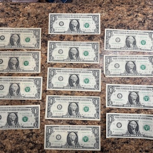 May include: A collection of fifteen United States one-dollar bills, each featuring a portrait of George Washington and the text "ONE DOLLAR". The bills are arranged on a brown surface.