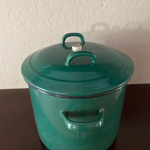 Op de afbeelding: Een grote, groene emaille pan met een deksel en drie handvatten. De pan staat op een donkerbruin oppervlak.