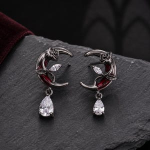 May include: A pair of crescent moon earrings in a dark gunmetal finish. Each earring features a small rose detail at the top, a deep red enamel accent along the curve, and a dangling clear pear-shaped crystal drop at the bottom of the moon.