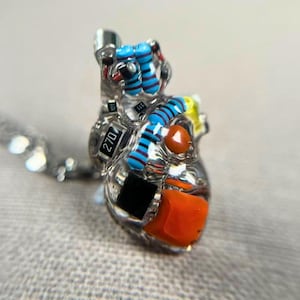 Puede incluir: Un colgante de corazón de resina transparente con componentes electrónicos integrados, incluyendo resistencias y condensadores. El colgante presenta una cadena plateada y está adornado con elementos naranjas, azules, negros y amarillos. El texto "270" es visible.