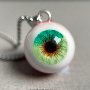 Puede incluir: Un colgante de collar con un dije de ojo detallado. El dije tiene un iris verde y marrón, sobre un fondo blanco, y está suspendido de una cadena plateada. El texto "Specter Bazaar" está en la parte inferior.