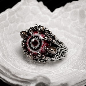 Puede incluir: Un anillo de metal plateado con un diseño central de ojo realista que presenta un iris rojo y una esclerótica blanca inyectada en sangre. La banda está elaborada con intrincadas puntas metálicas en forma de garra que rodean el ojo.