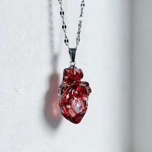 Puede incluir: Un collar con colgante en forma de corazón rojo y cadena plateada. El colgante es de un material rojo translúcido con detalles blancos, y la cadena tiene un diseño delicado. El texto "Specter Bazaar" está en la parte inferior.