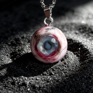 Puede incluir: Collar con colgante con diseño de ojo humano realista. El colgante es una esfera con iris azul, detalles rojos y blancos, suspendido de una cadena plateada. El texto "Specter Bazaar" está en la parte inferior.