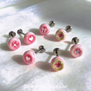 Puede incluir: Un conjunto de pendientes de botón rosas y blancos con varios diseños, incluyendo corazones, lazos y estrellas. Los pendientes tienen postes plateados y están dispuestos sobre una superficie blanca. Pequeños y delicados.