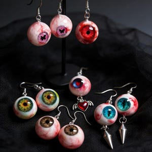 Puede incluir: Una colección de pendientes de ojo hechos a mano con varios colores de iris, incluyendo rosa, rojo, verde, azul y marrón. Algunos pendientes presentan detalles plateados y un corazón con alas. Los pendientes se exhiben sobre un fondo negro.