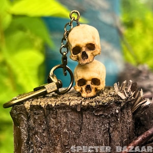 May include: A brass keychain with two small, stacked, bone-colored skull charms. The skulls have detailed features and are attached to a brass chain and a clasp.