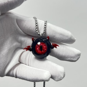 Puede incluir: Un colgante de ojo negro y rojo con cuernos y alas de diablo en una cadena plateada. El ojo es rojo con una pupila negra.