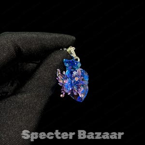 May include: A blue and purple pendant with a butterfly design. The pendant is made of a translucent material and has a silver-colored bail. The text "Specter Bazaar" is at the bottom of the image.