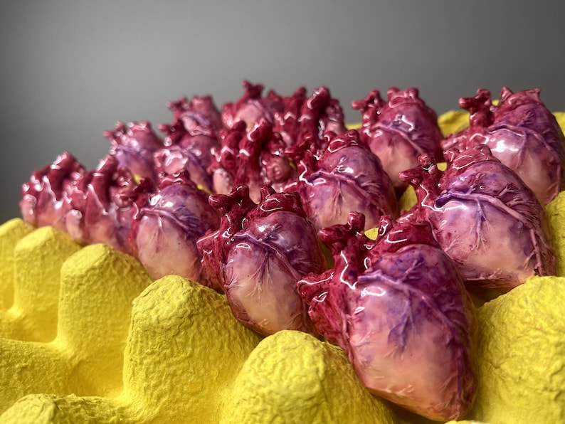 May include: A collection of small, anatomically detailed heart sculptures in shades of deep red, purple, and pale pink, each with a glossy finish, arranged within the individual molded compartments of a bright yellow cardboard egg carton.