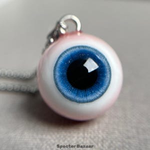 Puede incluir: Un collar con colgante de ojo. El colgante tiene un iris azul brillante y una pupila negra, sobre un fondo rosa y blanco. El collar cuelga de una cadena plateada. El texto "Specter Bazaar" está en la parte inferior.