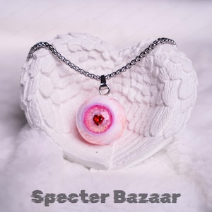 May include: A pendant necklace featuring a realistic eye design with a red heart-shaped pupil. The eye pendant is light pink and white, suspended from a silver chain. The necklace rests on a white, heart-shaped decorative piece. The text "Specter Bazaar" is at the bottom.