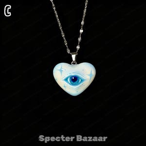 May include: A silver-toned chain necklace featuring a heart-shaped pendant. The pendant is light blue with a central dark blue eye graphic and four-pointed star accents. The letter C is visible in the top left corner and Specter Bazaar at the bottom.