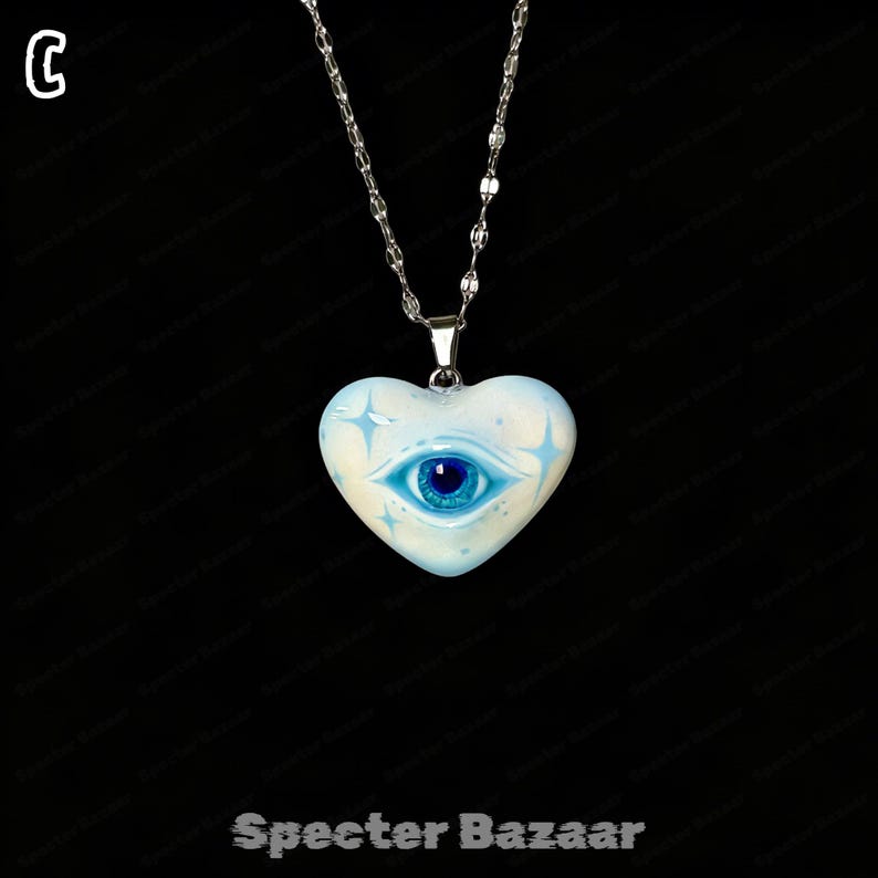 May include: A silver-toned chain necklace featuring a heart-shaped pendant. The pendant is light blue with a central dark blue eye graphic and four-pointed star accents. The letter C is visible in the top left corner and Specter Bazaar at the bottom.