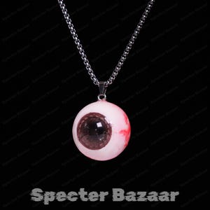 Puede incluir: Un collar con colgante de fantasía con un dije de ojo realista. El dije es de color rosa pálido con un iris marrón oscuro y venas rojas, suspendido de una cadena plateada. El texto "Specter Bazaar" está en la parte inferior.