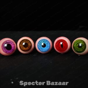 May include: Five prosthetic eyes with different iris colors: purple, brown, blue, red, and green. Each eye has a black pupil and is set against a black background. The text "Specter Bazaar" is at the bottom.