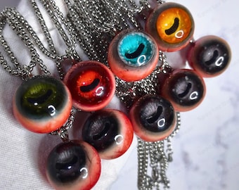 May include: A collection of handmade eye pendant necklaces. Each necklace features a round, glass-like pendant with a detailed eye design in various colors, including blue, green, red, and orange. The pendants are suspended from silver-colored chain necklaces.