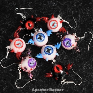 May include: A selection of handmade eyeball earrings. Each earring has a detailed eye design in various colours, including red, blue, and purple, with small wing accents. The earrings hang from silver-coloured ear wires. The text "Specter Bazaar" is at the bottom.