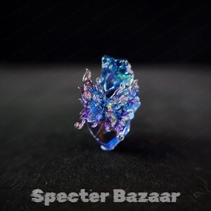 May include: A handcrafted, iridescent blue and purple glass ring with a floral design. The ring has a unique, artistic shape and is set against a dark background. The text "Specter Bazaar" is at the bottom.